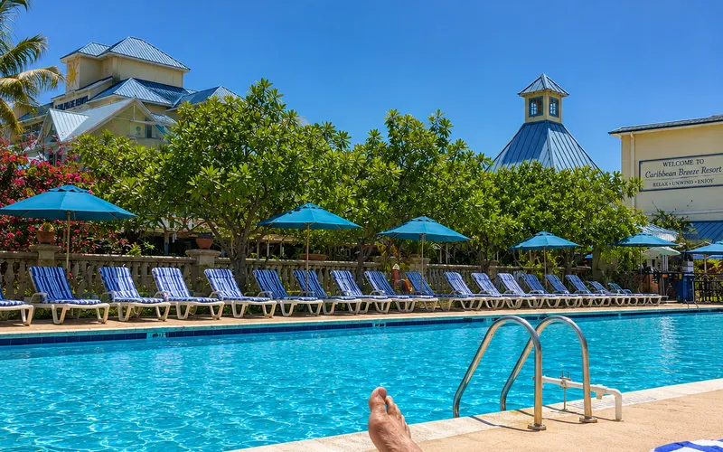 First-person view of feet up beside a Caribbean resort pool, striped loungers and blue umbrellas lining the water