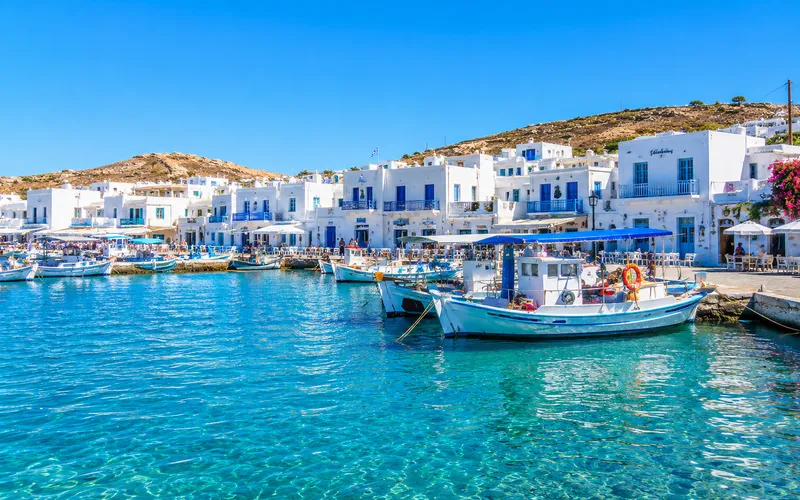 A Greek island harbor town with whitewashed buildings, blue shutters, and fishing boats moored in clear water