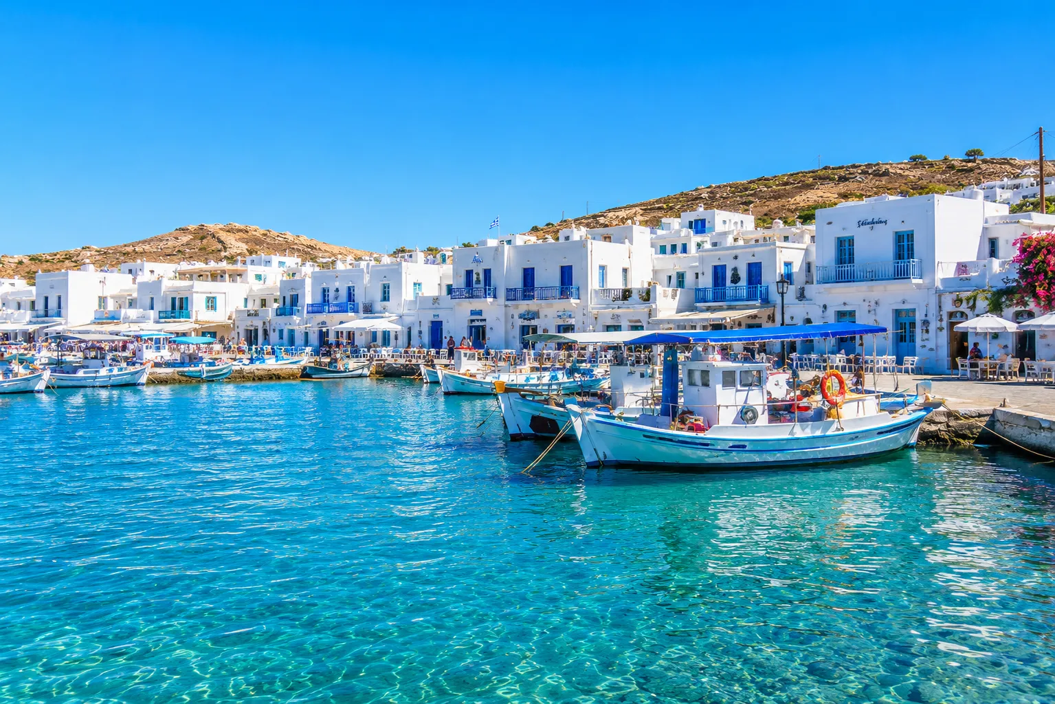 A Greek island harbor town with whitewashed buildings, blue shutters, and fishing boats moored in clear water