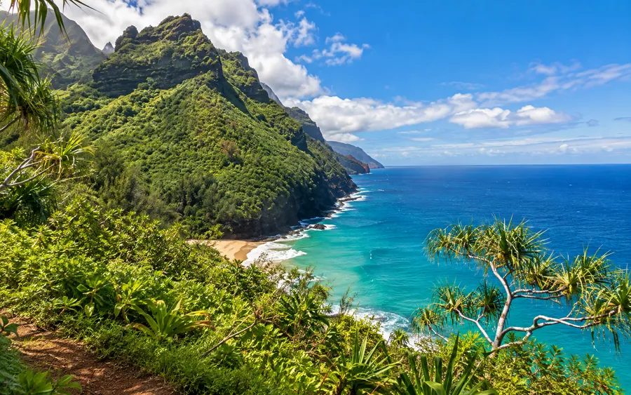 A secluded cove beach along Kauai's Nā Pali Coast, dramatic green fluted cliffs rising behind pale sand and turquoise water
