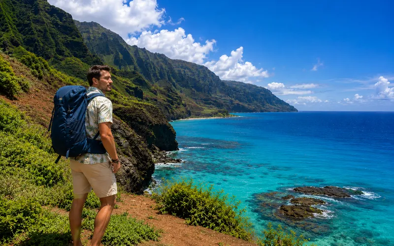 A hiker pausing on a coastal trail above Kauai's Nā Pali Coast, dramatic green cliffs and turquoise water below
