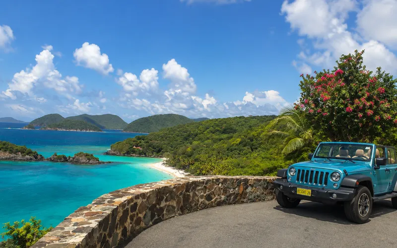 A rental Jeep parked at a stone-wall overlook above St. John, turquoise Trunk Bay and forested hills visible below