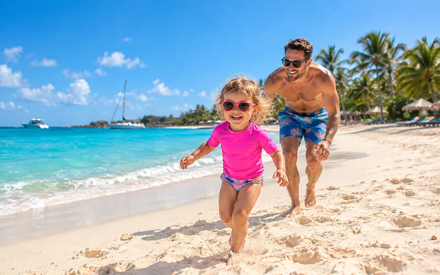 A toddler running along the shoreline with her dad close behind, calm turquoise Caribbean water and gentle waves
